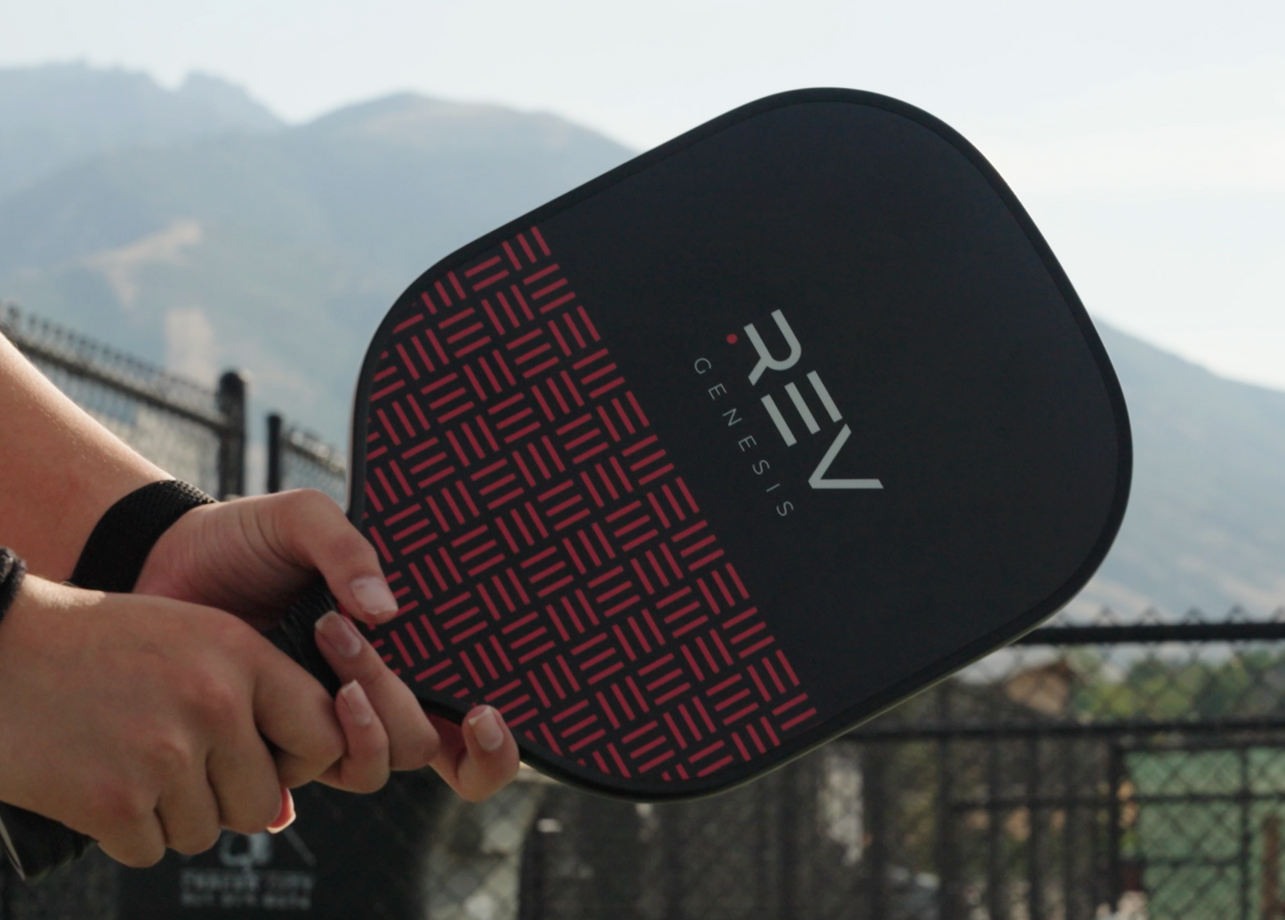 A close-up of someone holding a REV Pickleball GENESIS Pickleball Paddle Set with a red geometric design, ready to play outdoors. The background features mountains, a black chain-link fence, and clear skies.