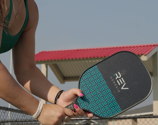 A person with pink nails and bracelets holds the REV Pickleball GENESIS Pickleball Paddle Set. Behind them, a fence and a red-roofed building stand beneath a clear sky.