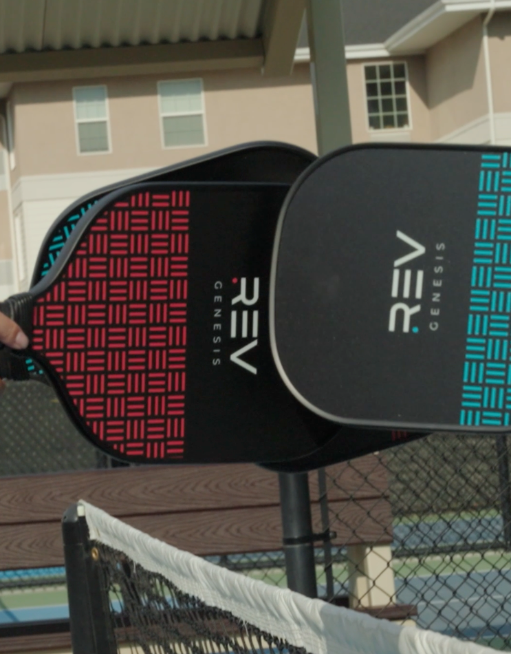 Close-up of two REV Pickleball GENESIS Pickleball Paddle Set paddles, one red and one blue, held above a net with a building and fence in the background—an ideal starter set for beginners.