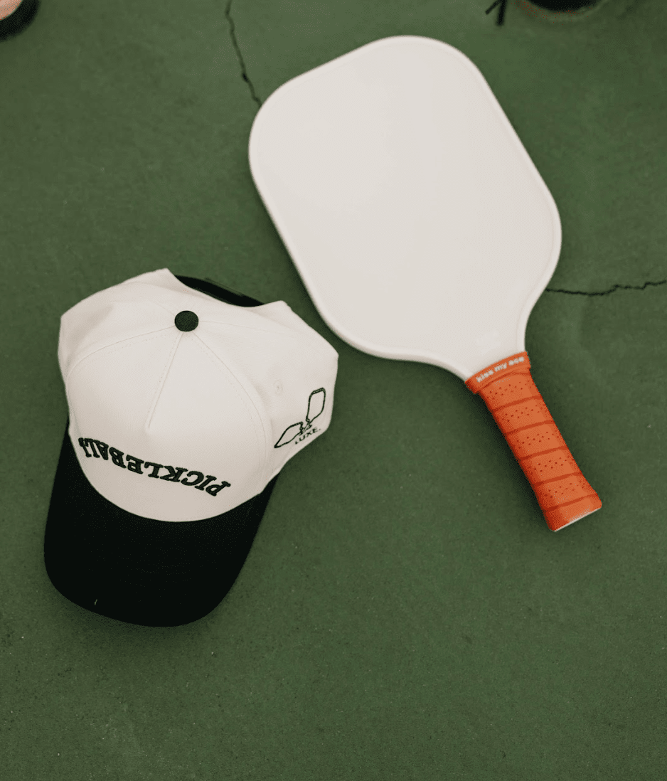 A white pickleball paddle with an orange grip rests on a green court next to the LUXE Pickleball Hat, a snapback featuring "PICKLEBALL" in reverse and a small paddle-and-ball design—stylish sun protection for every match.