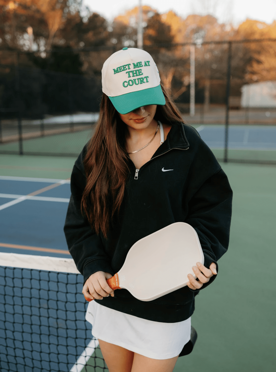 A woman stands on a pickleball court, wearing the LUXE Pickleball Meet Me At The Court Hat for sun protection, a black zip-up jacket, and white skirt. She holds a paddle as autumn trees and a tennis net appear in the background.