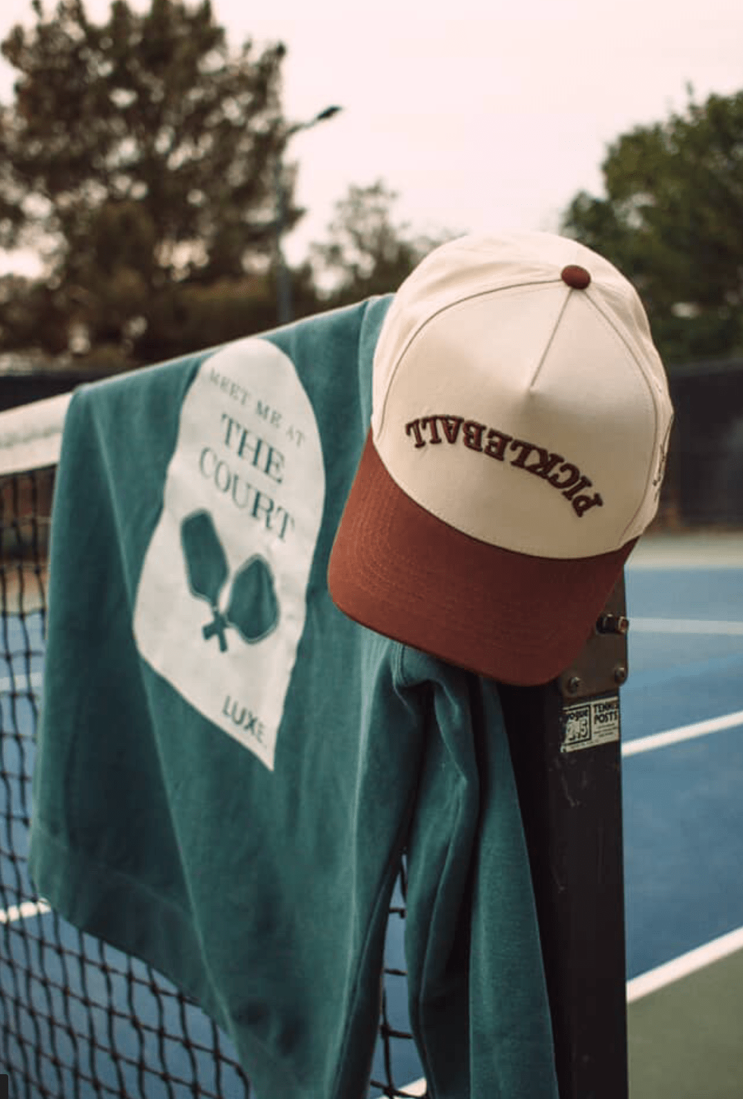 A LUXE Pickleball Hat with sun protection sits on a tennis net beside a green sweatshirt with paddle graphics and "MEET ME AT THE COURT LUXE," set on an outdoor court ringed by trees.