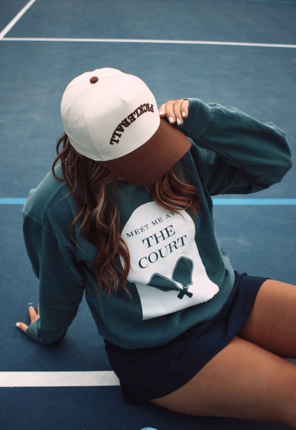 On a blue tennis court, someone relaxes courtside in a green "MEET ME AT THE COURT" sweatshirt, navy skirt, and the LUXE Pickleball Pickleball Hat snapback, which shades their face and offers sun protection.