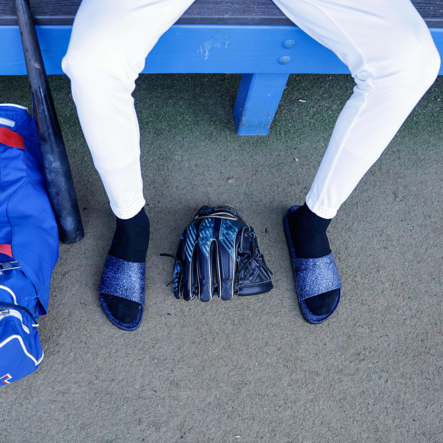 A person in white athletic pants sits on a bench with blue accents. On the ground are black socks, a bat, a black glove, and Blumaka FLEKS East Beach Slides from the Recovery Bundle next to a blue sports bag. The upper body is not visible.