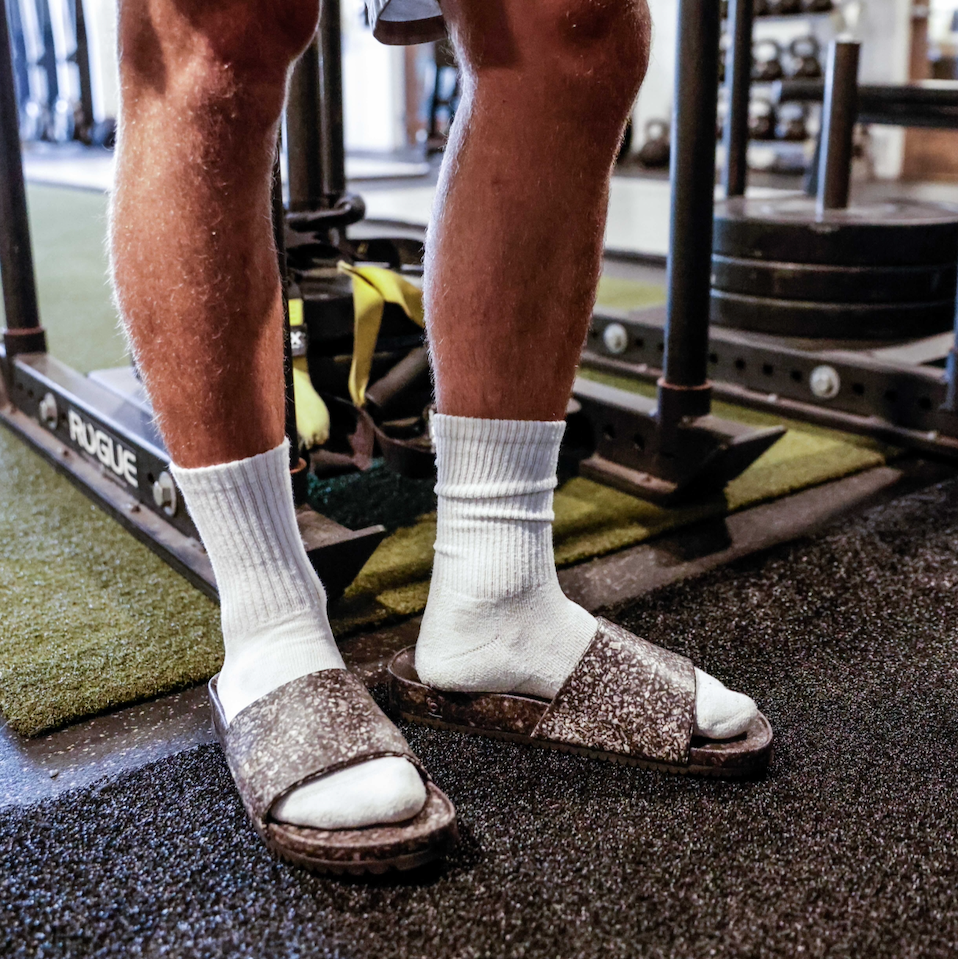 A person stands on a gym floor near weight plates and turf, wearing white socks and Blumaka Fleks® East Beach Slides. This fitness center scene suggests steps taken for athlete recovery or post-workout alignment support.