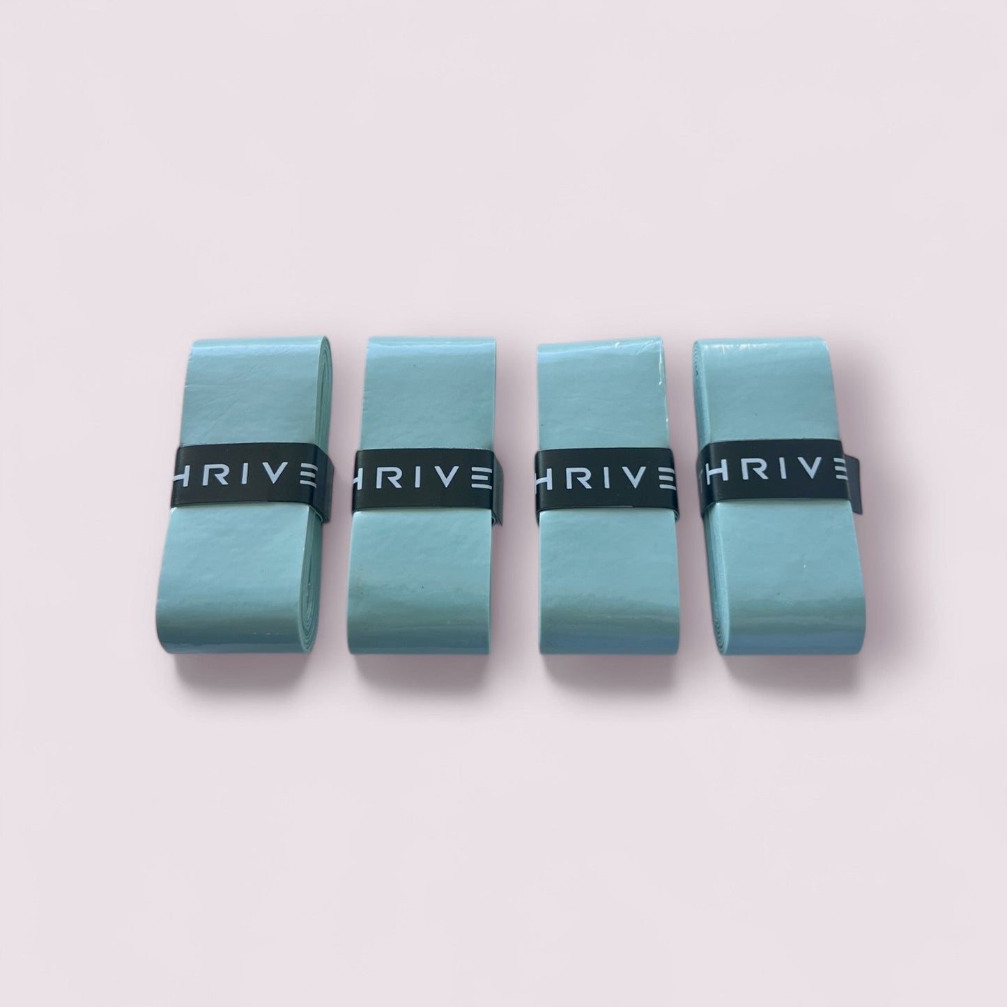 Four light blue resistance bands, each secured with a black Thrive Pickleball Overgrip (Super Tacky) featuring white "THRIVE" text, are neatly rolled and arranged side by side on a light pink background.
