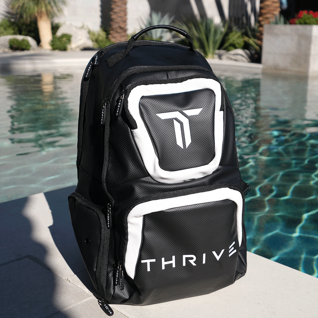 A black and white Thrive Pickleball Elite Backpack w/shoe bag, crafted from vegan leather, sits poolside in bright sunlight. Its bold logo faces the water, palm trees, and greenery while casting a sharp shadow on the concrete.