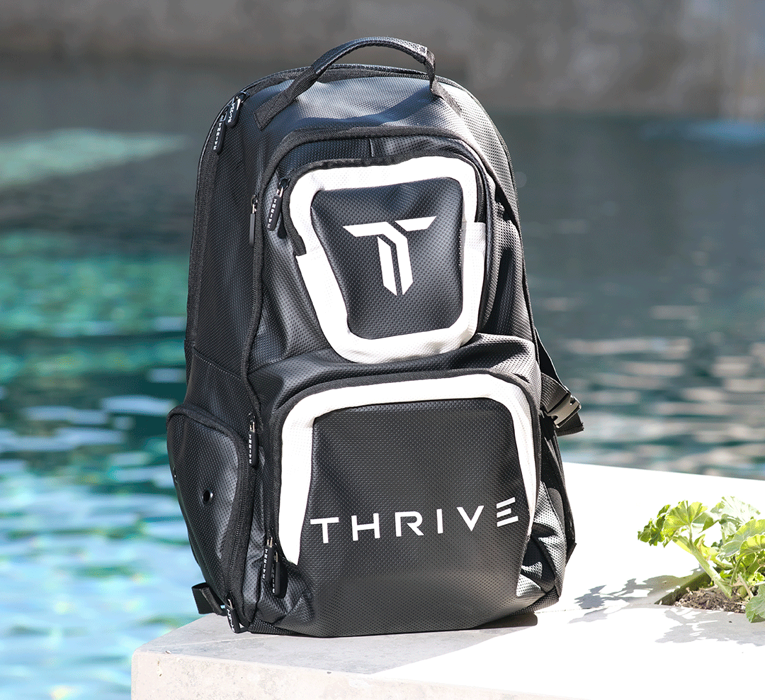 A black Elite Backpack w/shoe bag from Thrive Pickleball, featuring white accents and “THRIVE” on the front, made from vegan leather, sits on a concrete ledge near a pool with sparkling water and a green plant in view.