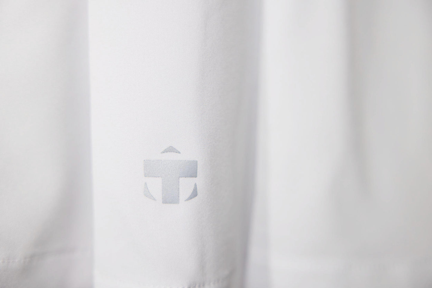 A close-up of the white TMPR Performance Mid-Rise Skort by TMPR Sports shows a subtle light grey logo with a stylized "T" and geometric shapes printed near the bottom left, set against a plain, evenly lit background.