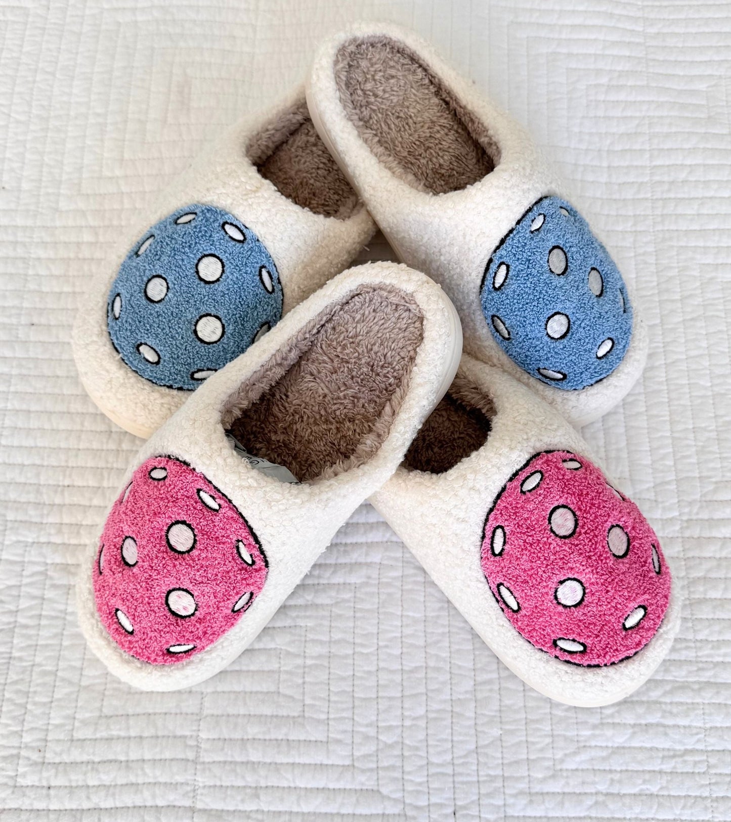 Fresh Pickle’s Pickleball Slippers feature cozy beige interiors and polka-dotted toe patches in blue or pink, making them perfect for lounging after a game. Resting on a textured white surface, these slippers combine comfort and playful style.