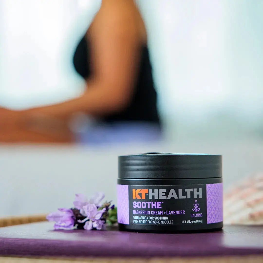 A jar of KT Tape KT Health Magnesium Creams with lavender rests beside fresh lavender, its label touting soothing benefits. In the background, a relaxed person embodies comfort and self-care.