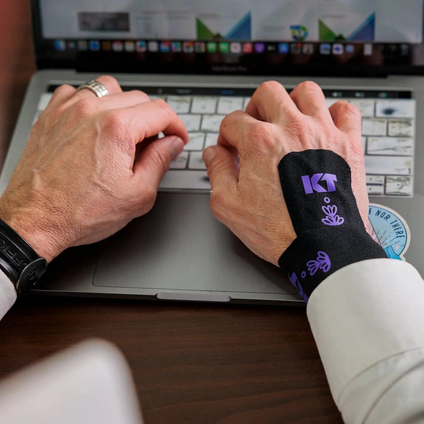 A person types on a laptop, wearing KT Tape Soothe by KT Tape with purple designs for pain relief on their right hand and wrist. The tape covers the back of the hand and wrist, with charts and tabs open on the laptop in the background.