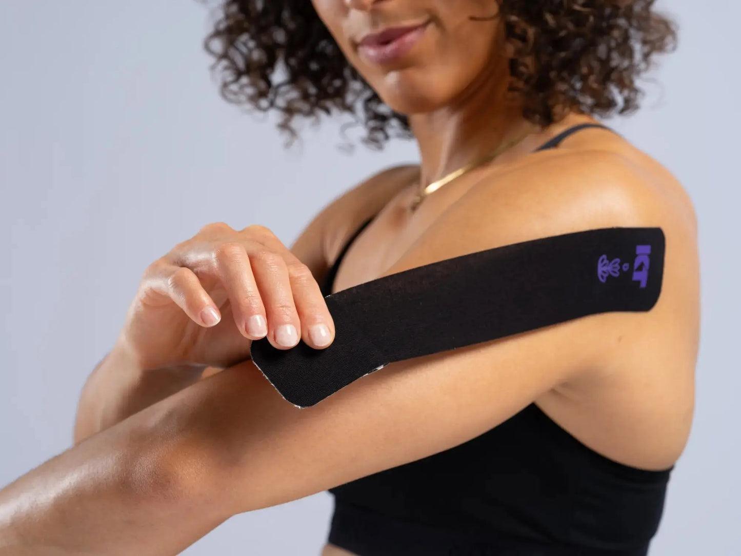 A woman with curly hair smiles slightly at the camera while applying a long black KT Tape Soothe strip by KT Tape to her upper arm for pain relief. The tape has a small purple logo and is set against a plain light gray background.