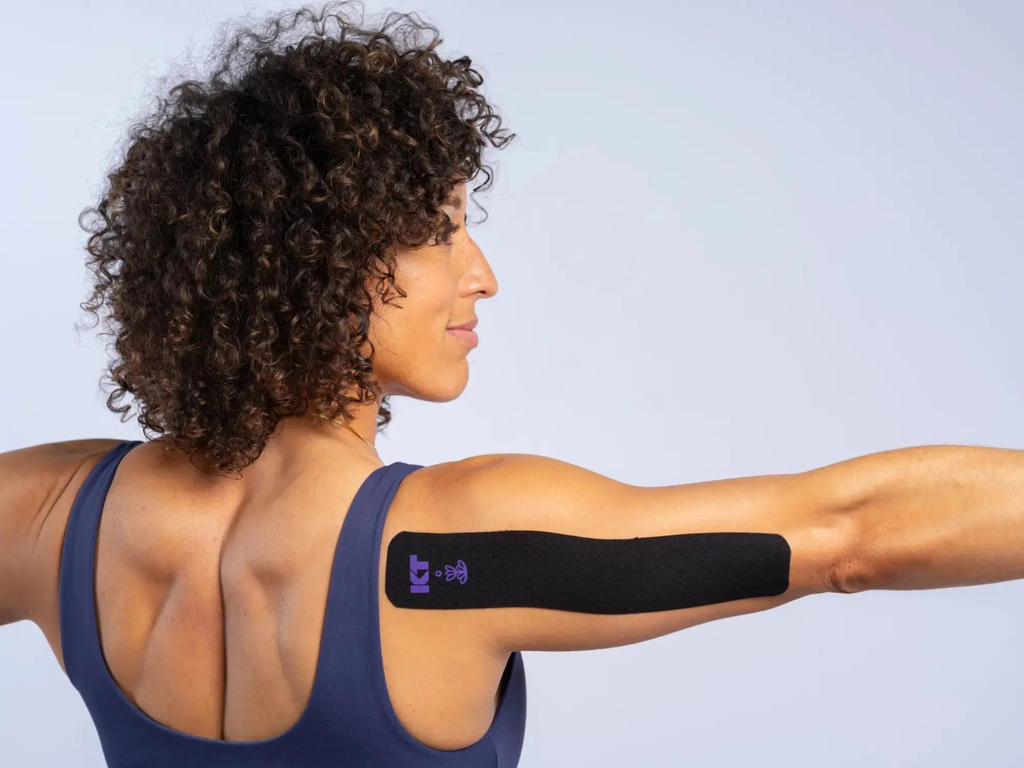 A woman with curly hair and a dark tank top stretches her arms out, displaying KT Tape Soothe by KT Tape—black kinesiology tape with a purple design—applied to her right upper arm for pain relief, set against a plain light background.