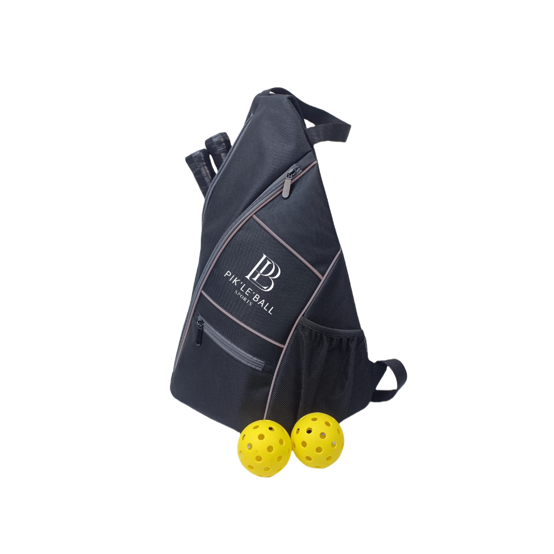 The Sporty Sling Grey Pickleball Bag by Pik'le'Ball Sports is a stylish, triangular bag with zipper pockets and branding on the front. Two paddles peek from the waterproof bag, with two yellow balls in front—must-have gear for every match.