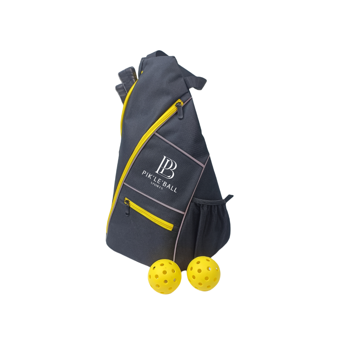 The Sporty Sling Yellow Pickleball Bag by Pik'le'Ball Sports stands upright, featuring dark gray fabric, yellow zipper accents, a white logo, and a right-side mesh pocket. Two yellow pickleballs are in front on a plain white background.