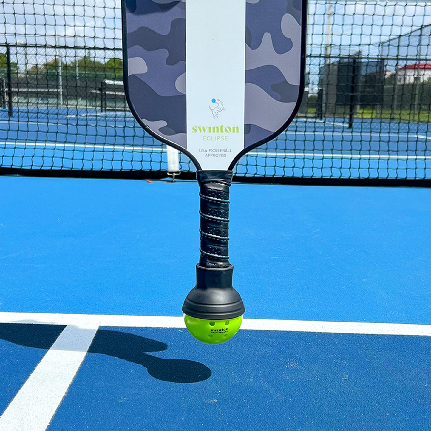 A Swinton Pickleball Suck Up paddle with a camouflage design stands upright on a blue court, featuring a green Suck Up accessory on the handle. A net and fence can be seen in the background.