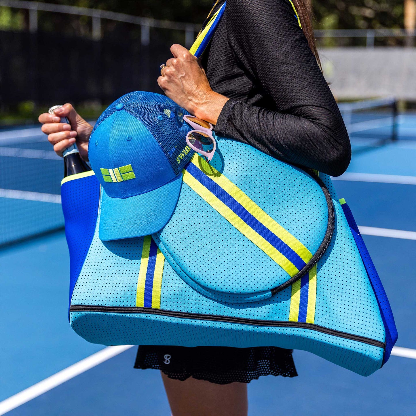 On a blue tennis court, someone carries the Swinton Pickleball XL Pickleball Tote Bag in blue, featuring green and yellow stripes. A blue cap and carabiner with keys hang from the handle, and a tennis racket is tucked inside.