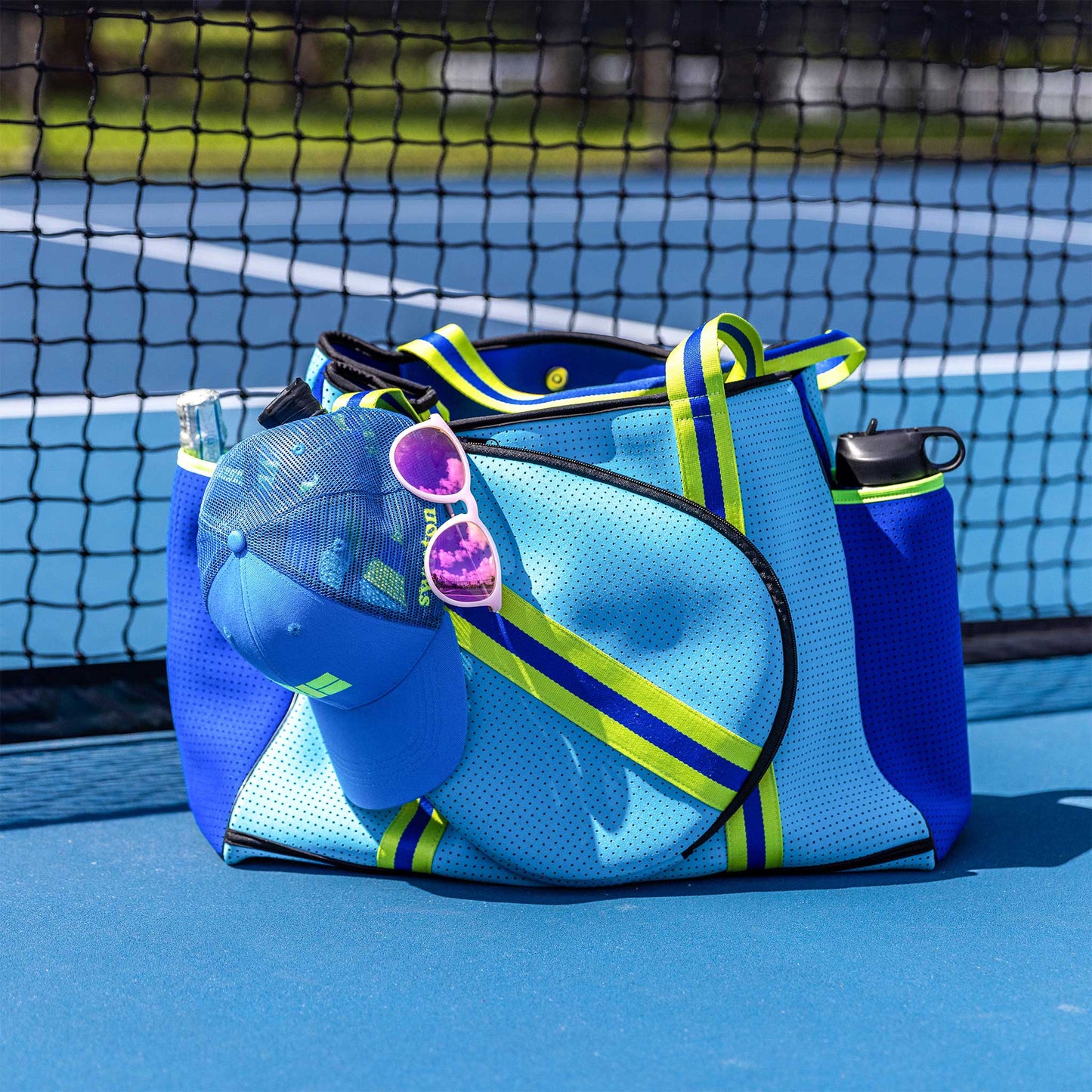 The Swinton Pickleball XL Pickleball Tote Bag - Blue rests against a net on a blue tennis court, holding a water bottle in its side pocket, with a blue cap and pink sunglasses hanging from it. The court surface and net are visible in the background.