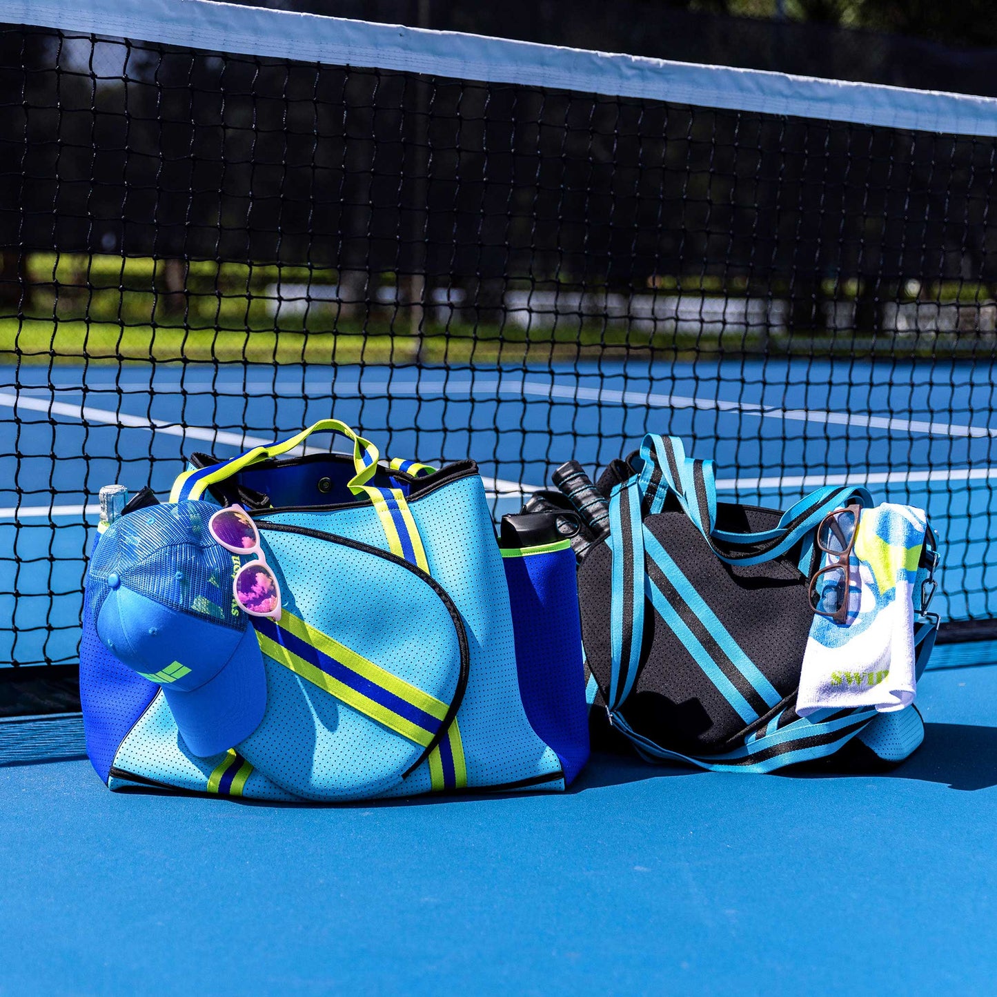 Two sporty bags rest on a blue tennis court: the Swinton Pickleball XL Pickleball Tote Bag - Black with yellow stripes holds a blue hat and purple sunglasses, while a sleek neoprene duffel sits by a bottle and palm tree towel in the sunlight.