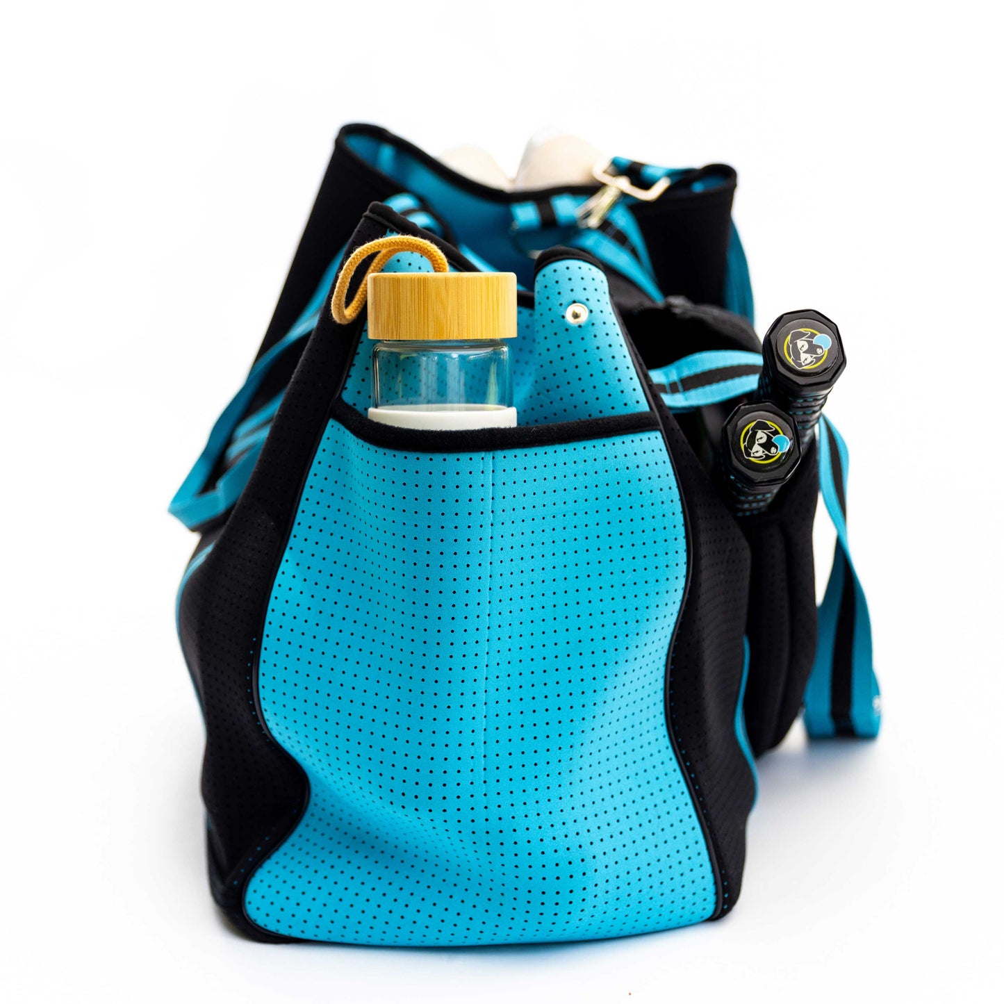 The Swinton Pickleball XL Pickleball Tote Bag - Black is pictured from the narrow end with perforated sides, a small clear container with a yellow lid in the outer pocket, and two black-handled rackets in the main compartment.