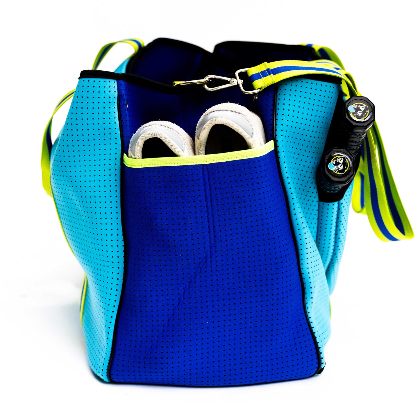 The Swinton Pickleball XL Pickleball Tote Bag - Blue, featuring turquoise neoprene with yellow accents and a striped yellow, blue, and green shoulder strap, sits on a white background with white sneakers partially visible in an outer pocket.