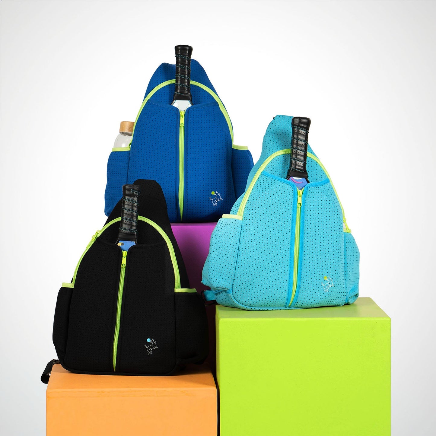 Three Swinton Pickleball Paddle Sling Bags in royal blue, black, and light blue with neon yellow zippers sit on pastel blocks. Each holds a paddle and water bottle, featuring a white animal logo on the pocket and durable neoprene accents.