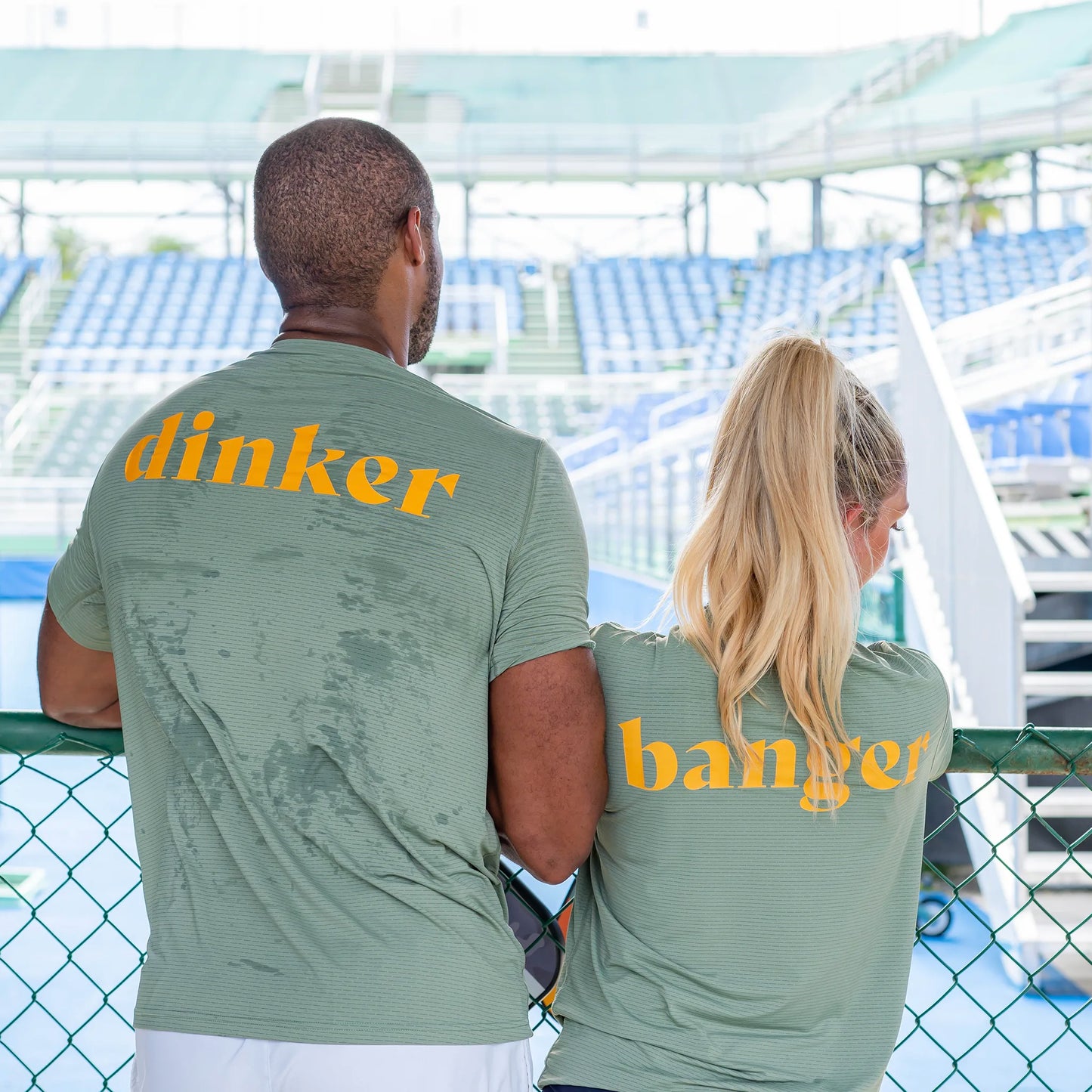 A man and woman, likely a doubles team, stand outdoors with their backs to the camera in matching Swinton Pickleball Women’s Banger LUX Performance Shirts, arms on a green fence and empty blue seats behind them.