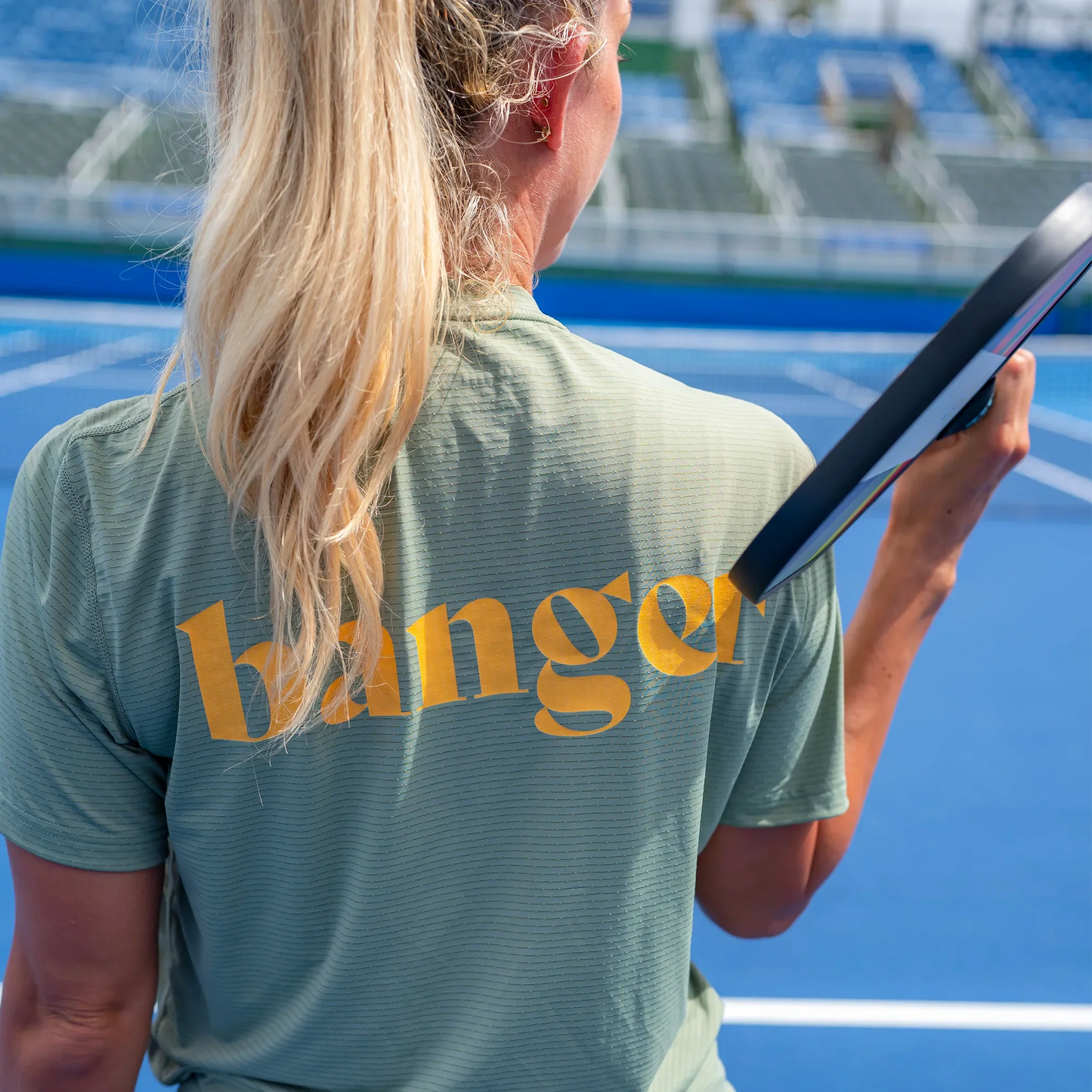 A woman with long blonde hair in a ponytail stands on a blue tennis court, wearing the Swinton Pickleball Women's Banger LUX Performance Shirt with "banger" in yellow across the back as she holds her racket.