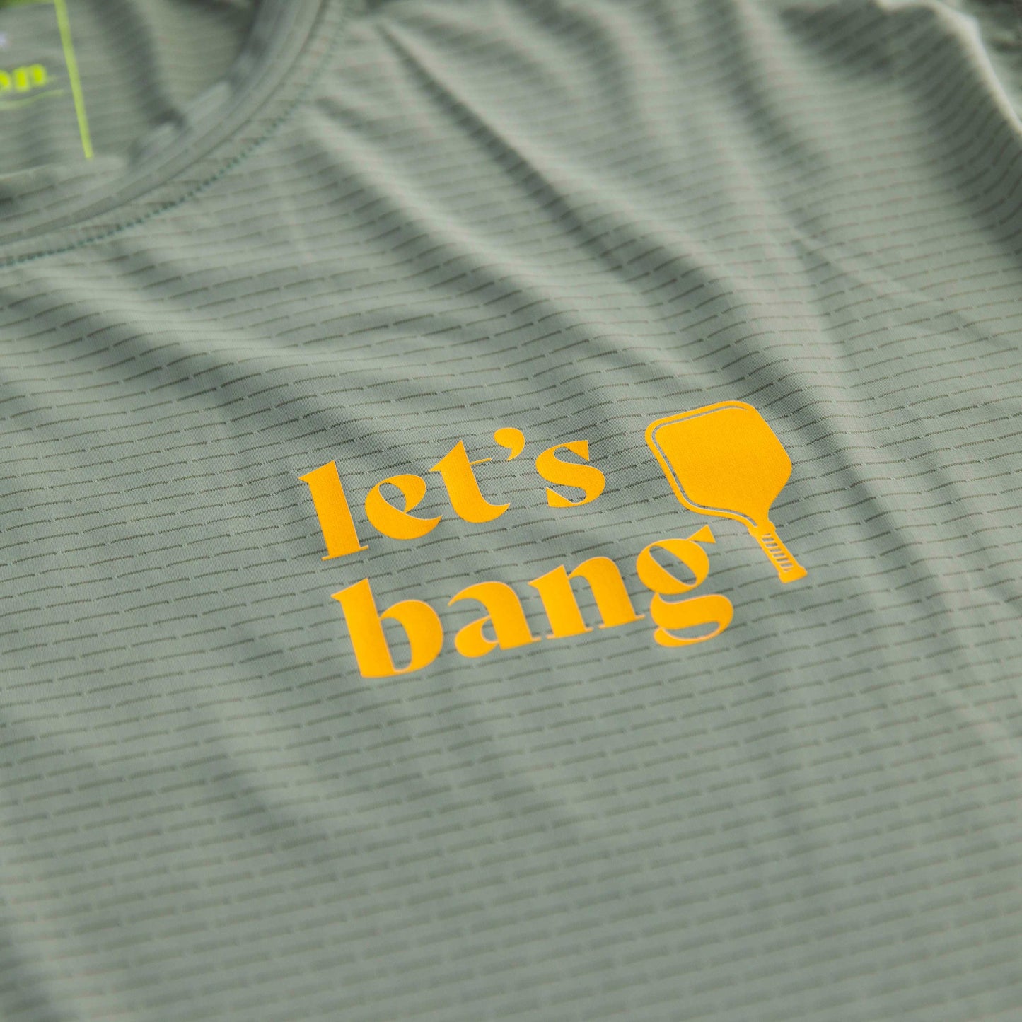 A close-up of the Women's Banger LUX Performance Shirt by Swinton Pickleball in light green with subtle horizontal stripes, made from performance material, features bold yellow “let’s bang” text beside a simple paddle illustration.
