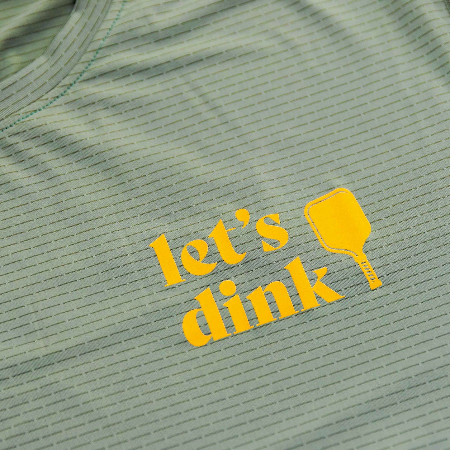 The Women's Dinker LUX Performance Shirt by Swinton Pickleball is a light green top with thin stripes, “lets dink” in bold yellow letters, and a yellow pickleball paddle graphic, crafted from soft performance material.