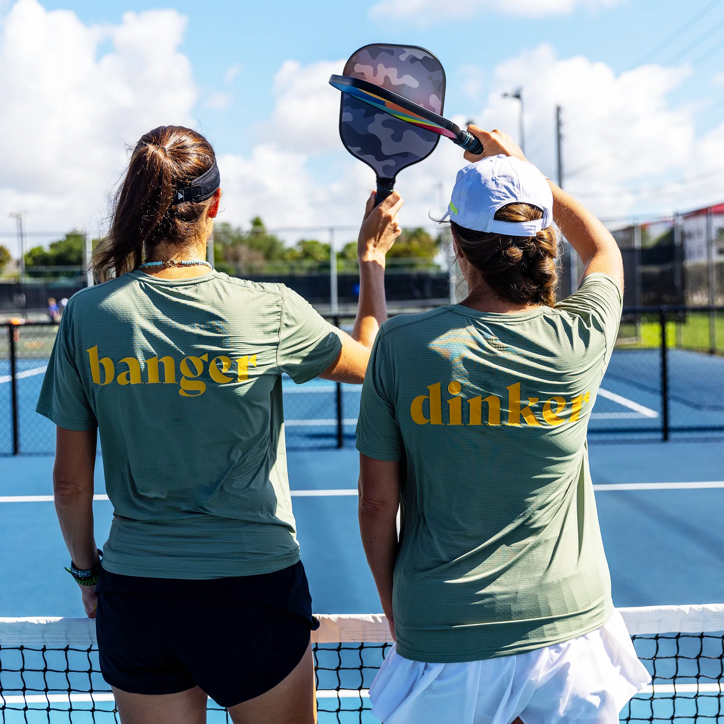 Two women play doubles on a pickleball court, one in a Swinton Pickleball Women's Dinker LUX Performance Shirt and hat, raising her paddle under the bright blue sky. The duo enjoys the game together, embodying playful team spirit.