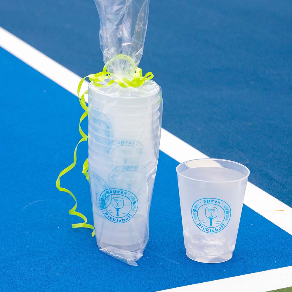 A stack of Swinton Pickleball’s Aprés Pickleball Reusable Party Cups, wrapped in plastic with yellow ribbon, sits on a blue court. One cup displays a blue martini logo and “apres pickleball”—the perfect gift for any player.