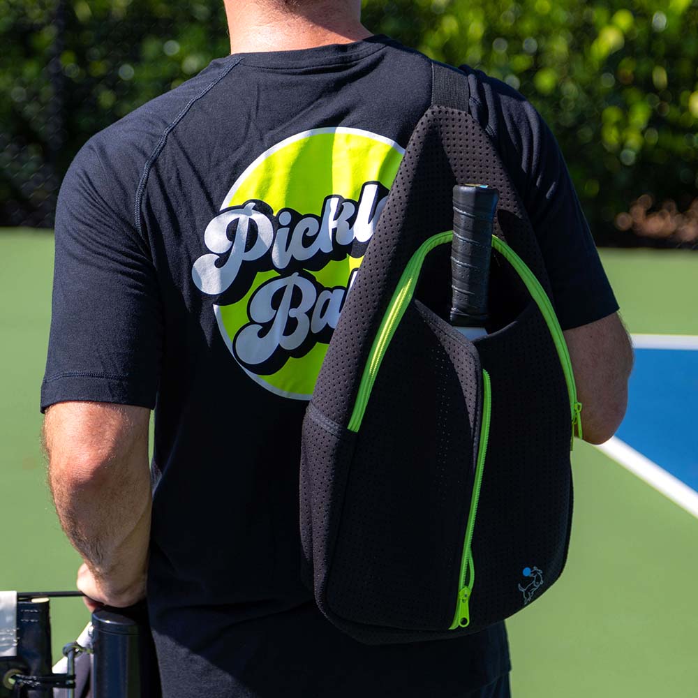 A person on an outdoor court carries the Swinton Pickleball Starter Bundle – Black Sling Bag Combo, featuring a pickleball paddle and accessories. Surrounded by greenery, it’s ideal gift inspiration for any pickleball enthusiast.