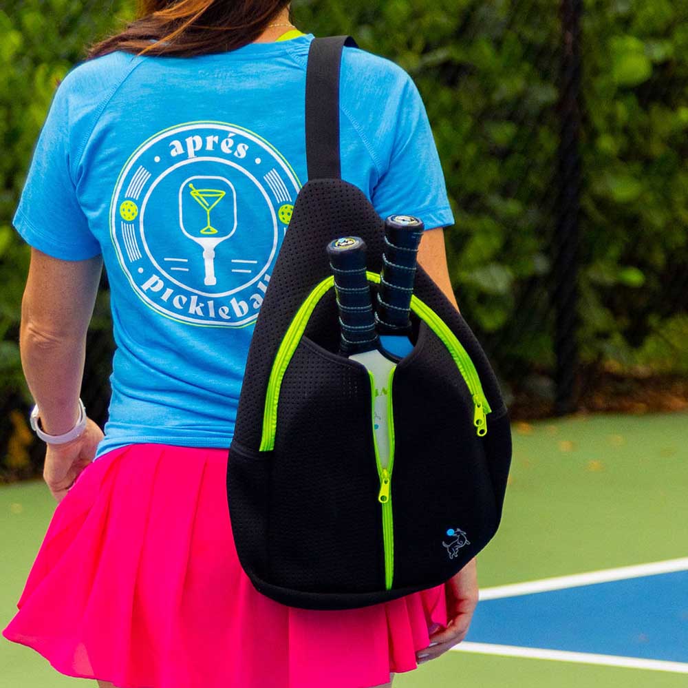 A woman in a blue “après pickleball” T-shirt and pink skirt stands on an outdoor court with the Swinton Pickleball Starter Bundle—Black Sling Bag Combo, the perfect gift and accessory for pickleball lovers. Green foliage frames the scene.