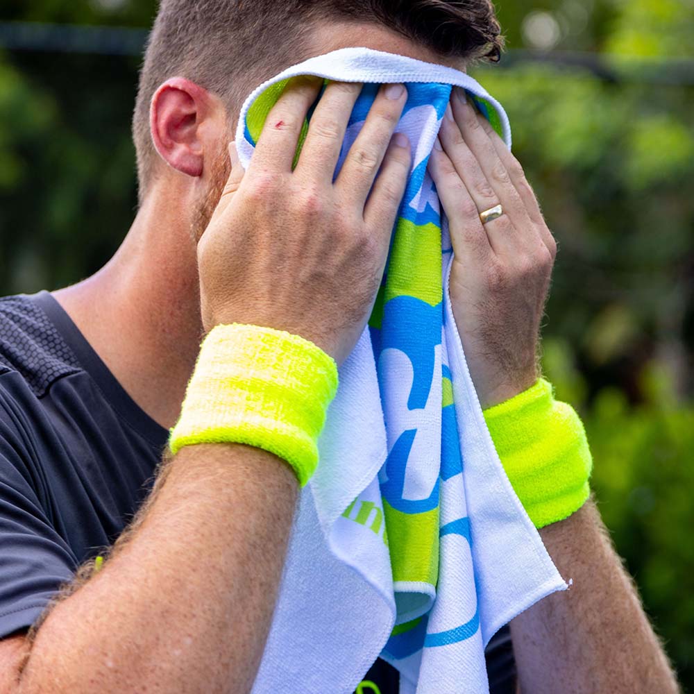A man in a dark shirt with yellow wristbands wipes his face with the Swinton Pickleball "Pickleball Sports Towel White." His wedding ring is visible, and the background shows blurry greenery. His face is completely covered by the towel.