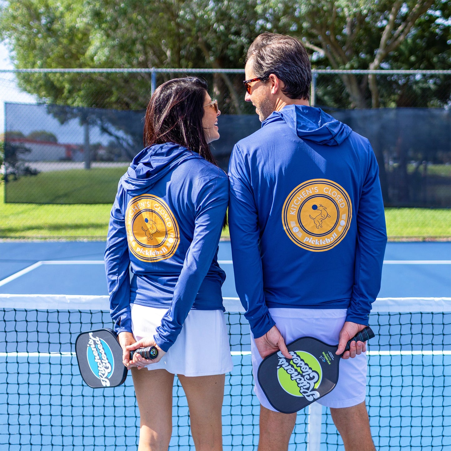 A man and woman stand side by side on a blue pickleball court, backs to the camera, wearing matching Swinton Pickleball Navy Zip-Front Sun Hoodies with a yellow “Pickleman’s Clubhouse” logo and holding paddles near the net.