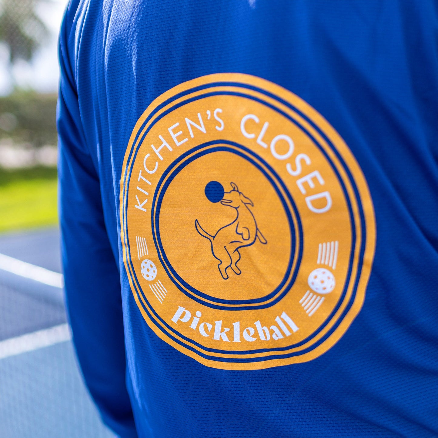A close-up of the back of a Swinton Pickleball Navy Zip-Front Sun Hoodie (UPF 50+) shows a circular yellow-orange design with a dog leaping at a pickleball, "KITCHENS CLOSED" text, and three small pickleball icons on a tennis court.
