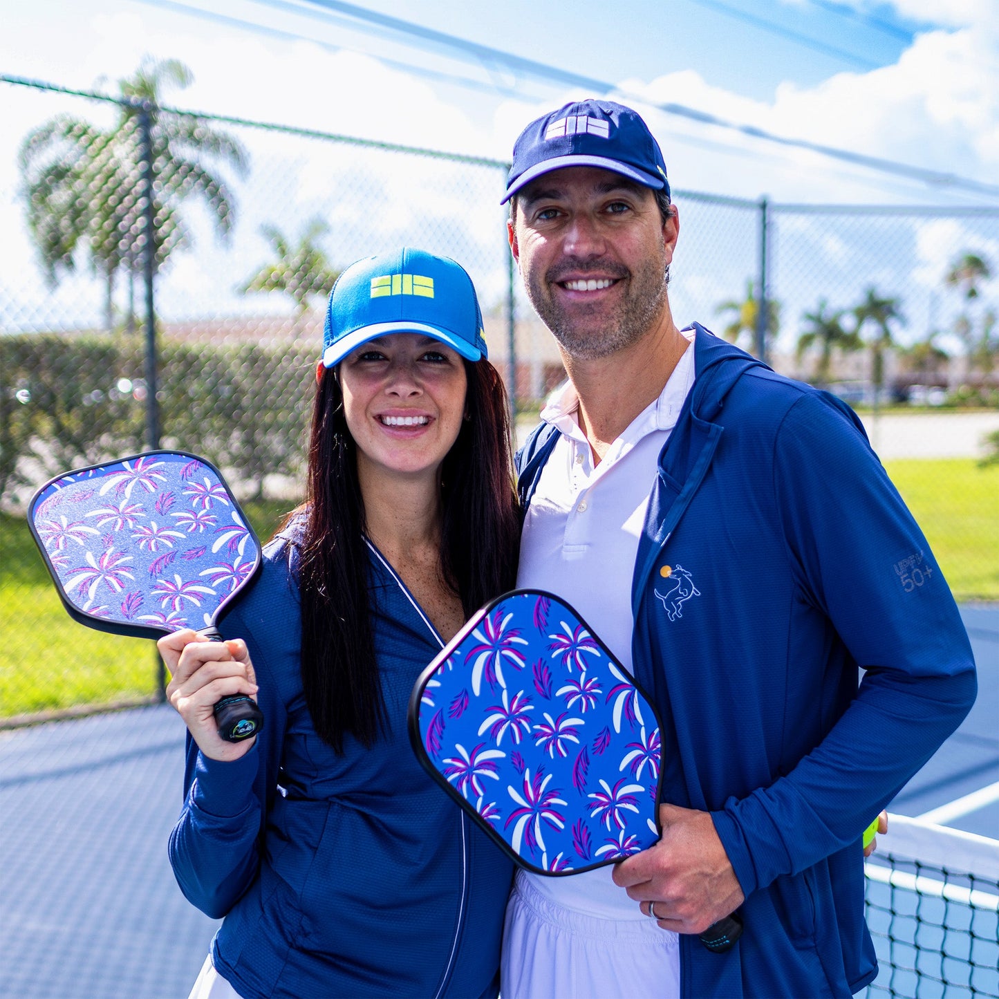 A smiling woman and man stand on an outdoor court, each holding a Swinton Pickleball Eclipse Island Vibes paddle featuring palm tree designs. Both wear blue jackets and baseball caps, with the net and palm trees in the sunny background.