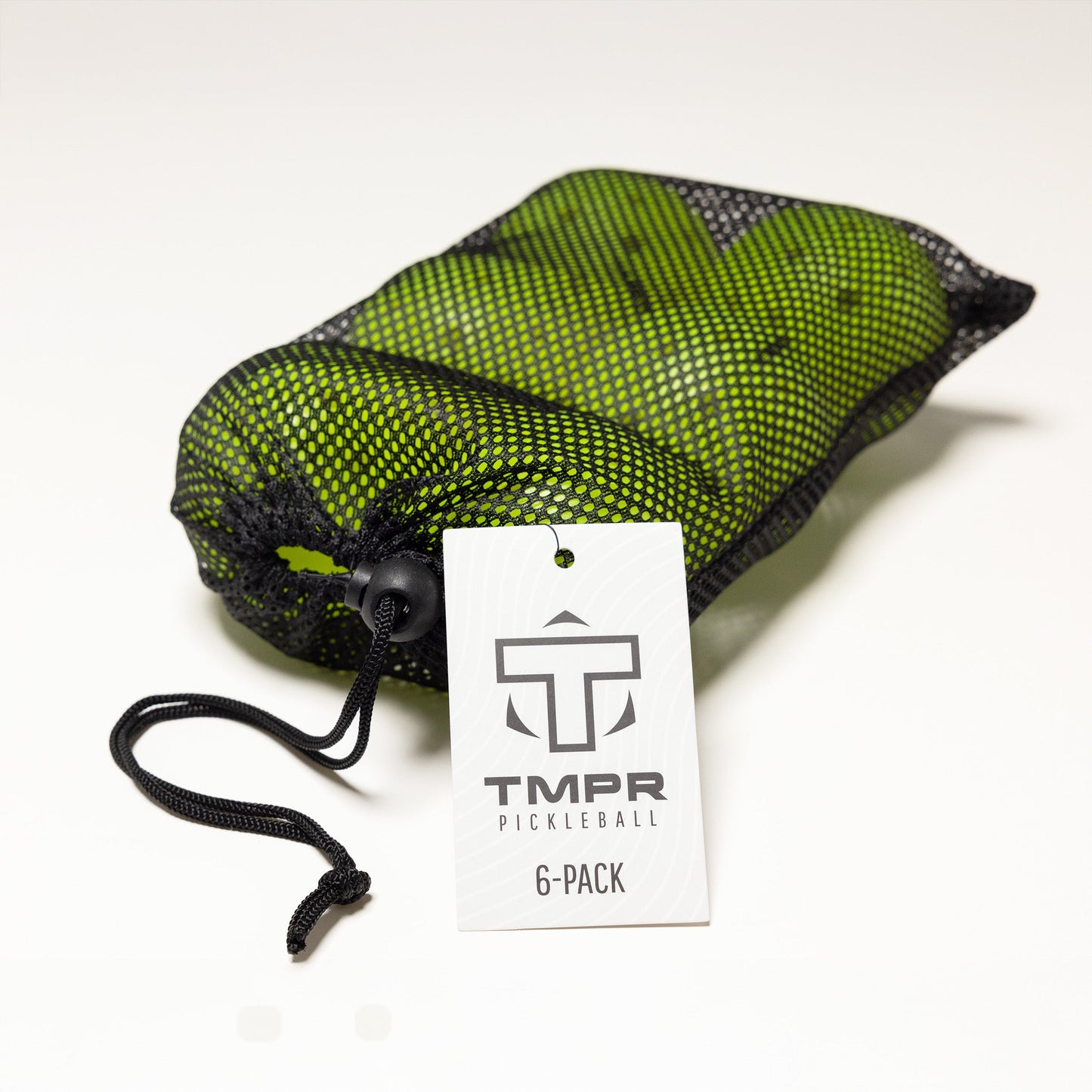 A black mesh drawstring bag holds six bright green TMPR Sports T-40 Outdoor Pickleballs. A white tag with the TMPR logo reads “T-40 Outdoor Pickleball, 6-Pack.” The bag rests on a plain white surface.