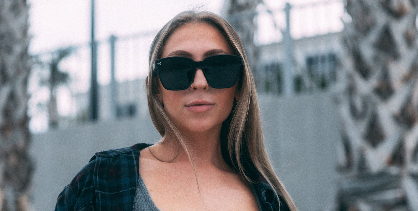 A woman with long, straight blonde hair wears Neven Eyewear Atlantis sunglasses with polarized UV400 protection. She stands outdoors by palm trees and a metal fence in a plaid shirt over a gray top, looking confidently at the camera.