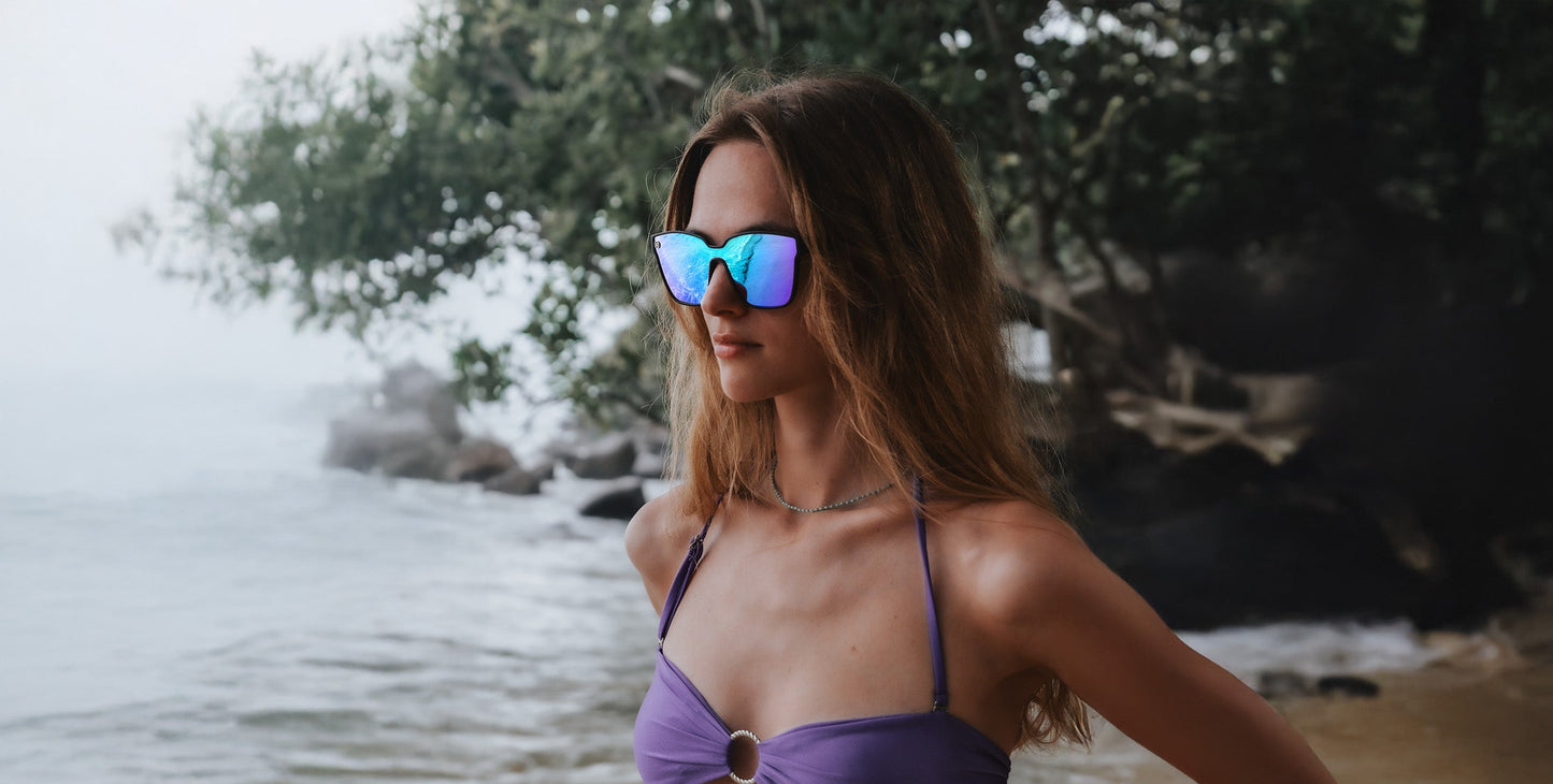 A woman wearing Neven Eyewear's Curiosity sunglasses with a matte black frame and a lilac swimsuit stands on a beach, gazing to the side. Her long, wavy light brown hair flows as green trees, rocks, and mist fill the background.