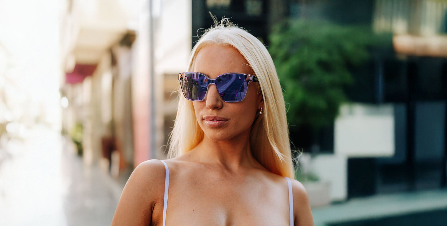 A woman with long platinum blonde hair wears Neven Eyewear's Discovery sunglasses with polarized UV400 protection and a white tank top, standing outdoors on a sunlit street with blurred greenery and modern buildings in the background.