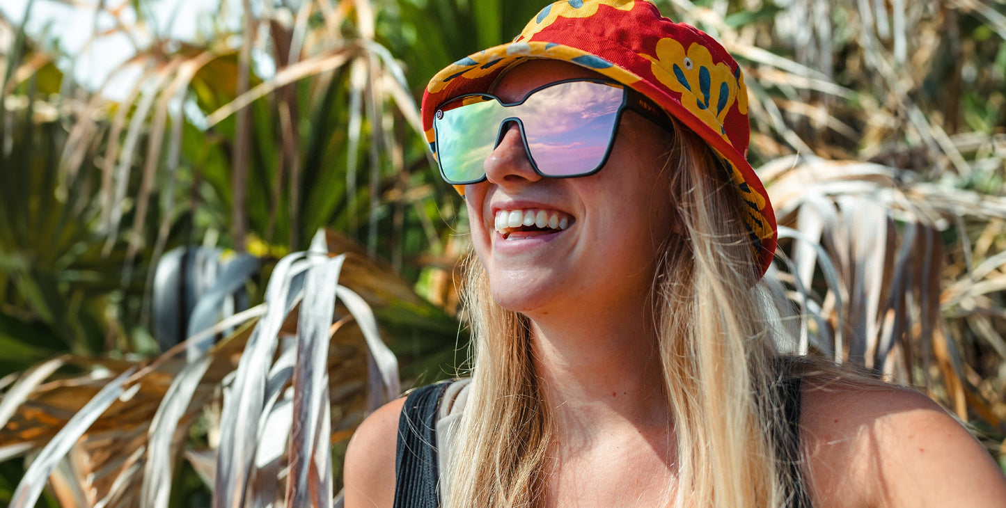 A smiling woman with long blond hair wears a colorful bucket hat and Neven Eyewear Endeavour sunglasses, which have polarized UV400 protection and large mirrored lenses reflecting a pink-blue sky. She stands outdoors amid blurred green and brown foliage.