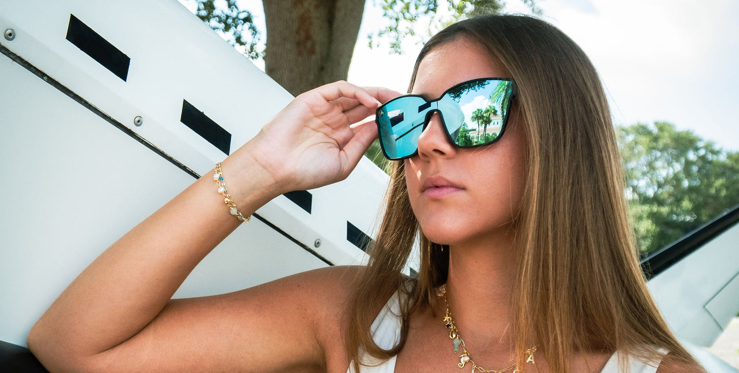 A woman with straight brown hair adjusts her Neven Eyewear Perseverance sunglasses, which are space-inspired, reflective blue with UV400 protection. She wears a white top and gold jewelry outdoors, with trees and a white vehicle in the background.