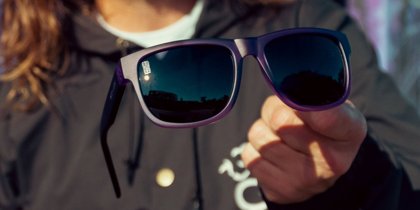 A person with long hair in a dark jacket holds Neven Eyewear Ella rectangular sunglasses toward the camera. The black lenses reflect a sunny scene, while the person's face is out of focus in the background.