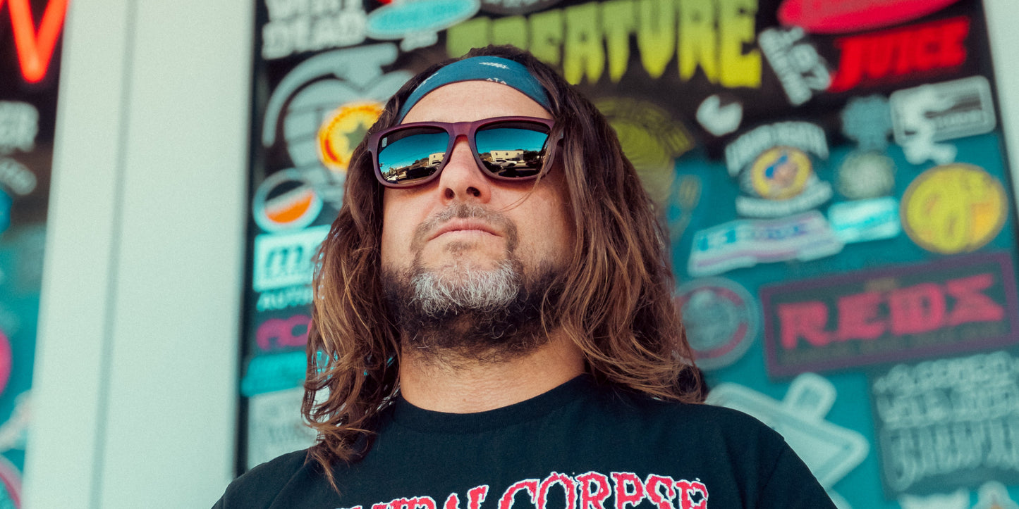 A man with long brown hair and a beard wears Neven Eyewear's Plasma sunglasses and a blue headband. He stands before a glass window covered in colorful stickers, gazing confidently ahead in a black T-shirt with a red and white design.