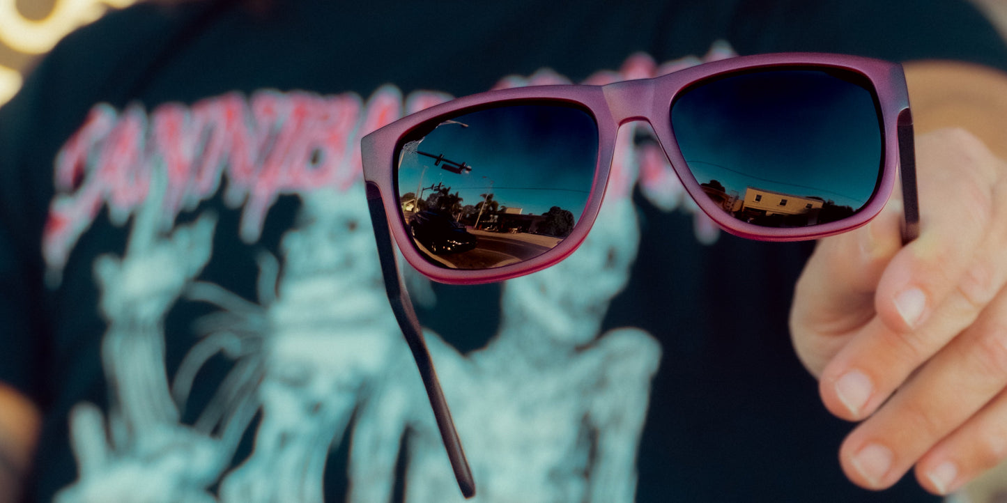 A person holds out Neven Eyewear Plasma sunglasses with purple rims and polarized lenses reflecting a clear sky, a small airplane, power lines, trees, and buildings. They're wearing a dark T-shirt with colorful abstract graphics and blurred text.
