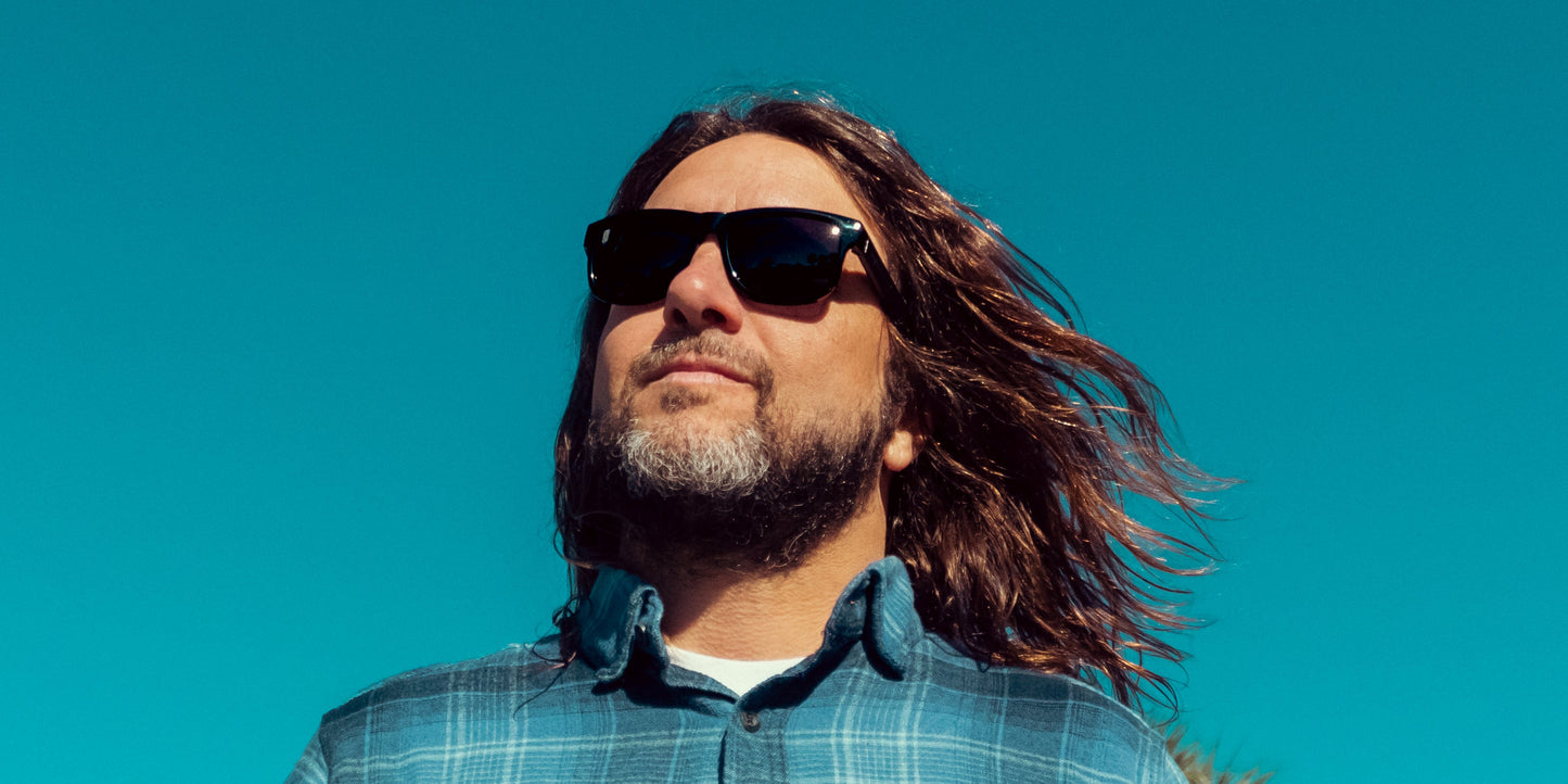 A man with long brown hair and a beard wears Neven Eyewear Sade rectangular sunglasses and a blue plaid shirt outdoors against a bright sky. His hair blows in the wind as he looks ahead with a calm, confident expression.