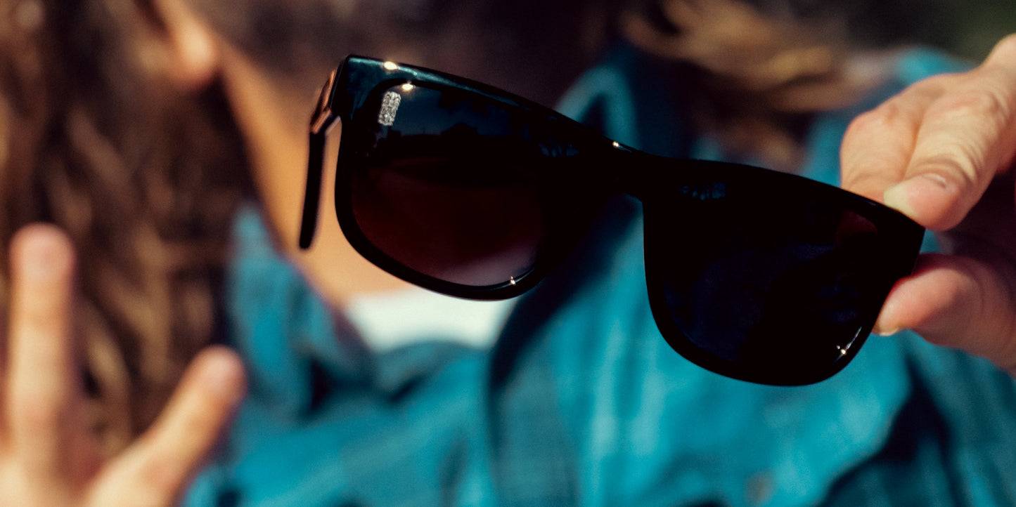 A person in a blue plaid shirt holds out a pair of Sade polarized sunglasses by Neven Eyewear toward the camera, with the sharply focused sunglasses in the foreground and the blurred individual in the background.